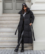 Black long puffer coat with hood up, tie belt at waist and quilted front