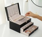 Black leather jewelry box open with layered trays and a hand arranging necklaces