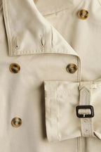 Light beige trench coat detail: gold-tone buttons, pocket flap, and waist belt buckle