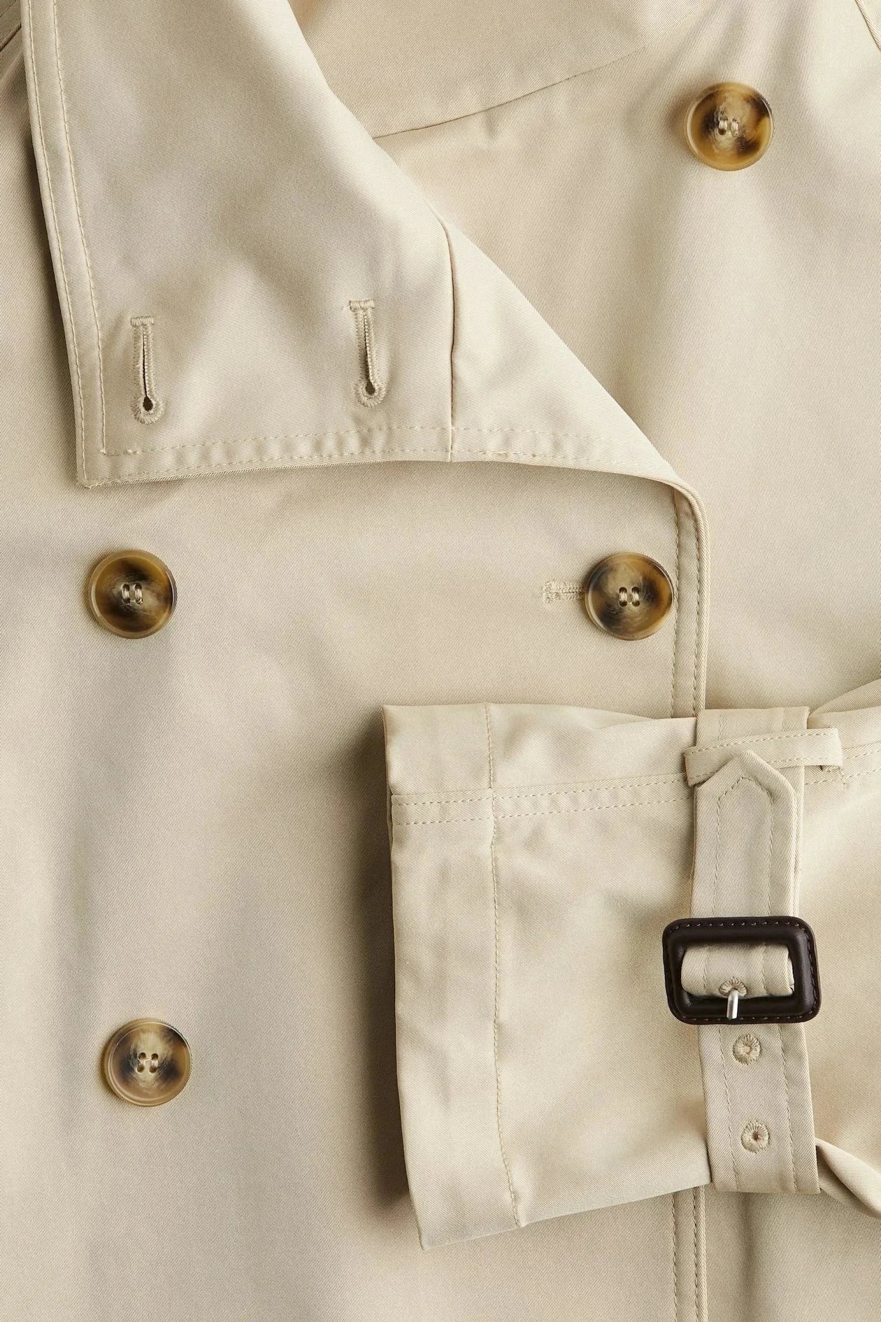 Light beige trench coat detail: gold-tone buttons, pocket flap, and waist belt buckle